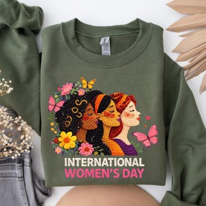 May include: Olive green sweatshirt featuring an embroidered design of three women with flowers and butterflies. The text "INTERNATIONAL WOMEN'S DAY" is displayed in pink and white. The sweatshirt has long sleeves and a crew neck.