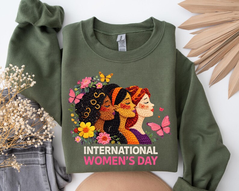 May include: Olive green sweatshirt featuring an embroidered design of three women with flowers and butterflies. The text "INTERNATIONAL WOMEN'S DAY" is displayed in pink and white. The sweatshirt has long sleeves and a crew neck.