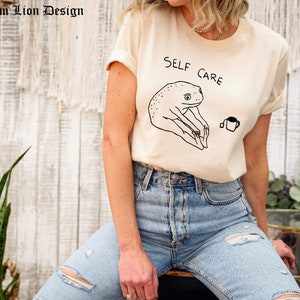 May include: A beige t-shirt with a black line drawing of a frog holding a cup of tea. The text "SELF CARE" is written above the frog.