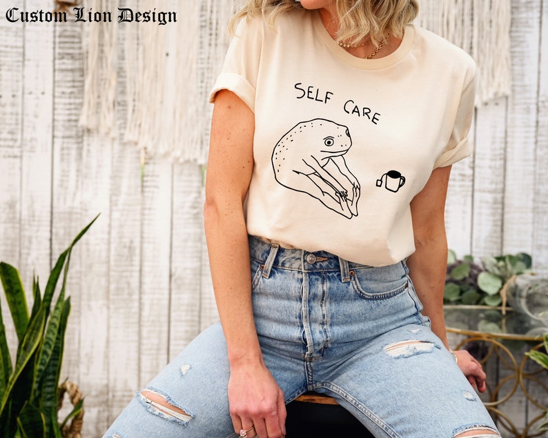May include: A beige t-shirt with a black line drawing of a frog holding a cup of tea. The text "SELF CARE" is written above the frog.