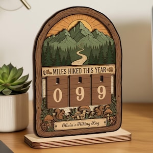 May include: Wooden hiking log with a mountain scene, displaying "MILES HIKED THIS YEAR" and the number "099". The log features a forest design with squirrels and mushrooms, and the text "Olivia's Hiking Log".