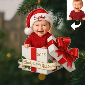 Puede incluir: Adorno navideño con un bebé que lleva un gorro de Papá Noel con el nombre "Sophia". El bebé está dentro de una caja de regalo blanca con una cinta roja, el texto "Baby's 1st Christmas" y el año "2025".