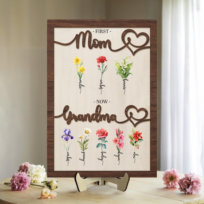 May include: A wooden sign with the words "First Mom" and "Now Grandma" in a cursive font. The sign is decorated with a heart and flowers. The flowers are labeled with names, such as Millie, Bonnie, and Jenny.