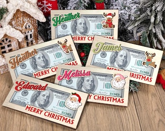 Könnte beinhalten: Holztafeln, die 100-Dollar-Scheinen ähneln, personalisiert mit Namen wie "Heather" und "James". Jede Tafel hat ein festliches Weihnachtsthema mit dem Text "Merry Christmas" und Weihnachtsaufklebern wie Santa und Rentieren.