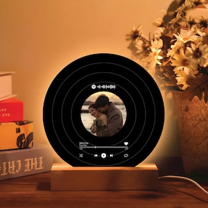 May include: A black circular lamp with a wooden base. The lamp has a built-in photo frame and a music player with a heart icon, play and pause buttons, and a progress bar. The text "Marry You" and "Bruno Mars" is displayed on the lamp.