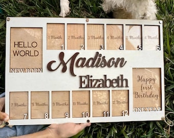May include: White wooden photo frame with 12 slots for pictures. The frame is decorated with the words "Hello World", "New Born", "Madison Elizabeth", "Happy First Birthday", and "One Year". Each slot is labeled with a month from 1 to 12.