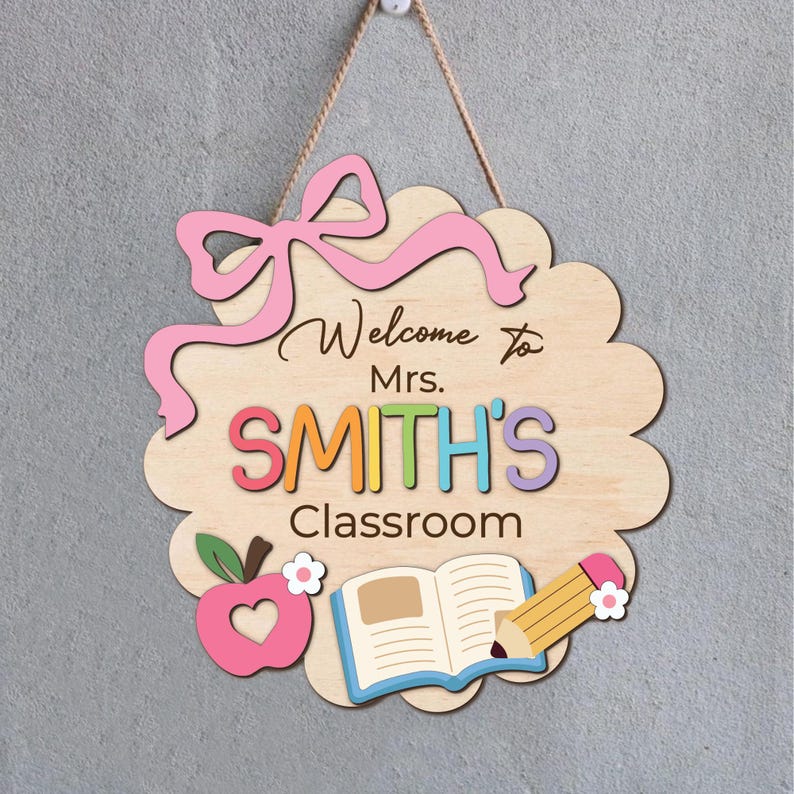 K&ouml;nnte beinhalten: Ein Schild aus Holz f&uuml;r eine Klassent&uuml;r mit einer rosa Schleife, dem Text "Willkommen in der Klasse von Frau SMITH", einem Apfel, einem ge&ouml;ffneten Buch und einem Bleistift.