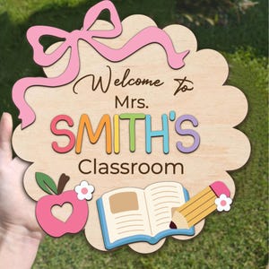 K&ouml;nnte beinhalten: Holzschild f&uuml;r eine Klassent&uuml;r mit einer rosa Schleife, dem Text "Willkommen in der Klasse von Frau SMITH", einem Apfel mit einem Herzen, einem Buch und einem Bleistift.