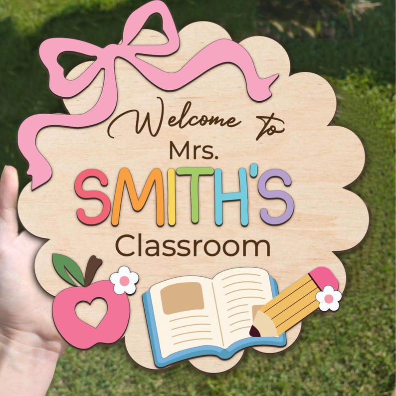 K&ouml;nnte beinhalten: Holzschild f&uuml;r eine Klassent&uuml;r mit einer rosa Schleife, dem Text "Willkommen in der Klasse von Frau SMITH", einem Apfel mit einem Herzen, einem Buch und einem Bleistift.