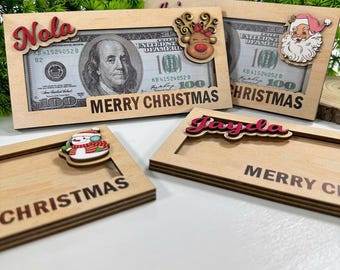 Könnte beinhalten: Weihnachtsrahmen aus Holz, die für eine 100-Euro-Note konzipiert sind. Jeder Rahmen hat einen Namen, den Text „Frohe Weihnachten“ und eine festliche Verzierung wie ein Rentier, einen Weihnachtsmann oder einen Schneemann. Die Rahmen sind aus hellem Holz.