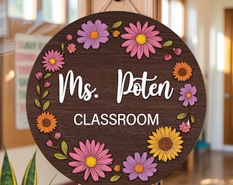 Pode incluir: Uma placa de sala de aula redonda de madeira com fundo marrom escuro. Decorada com flores coloridas e o texto "Ms. Poten CLASSROOM" em branco. A placa está pendurada em uma porta.