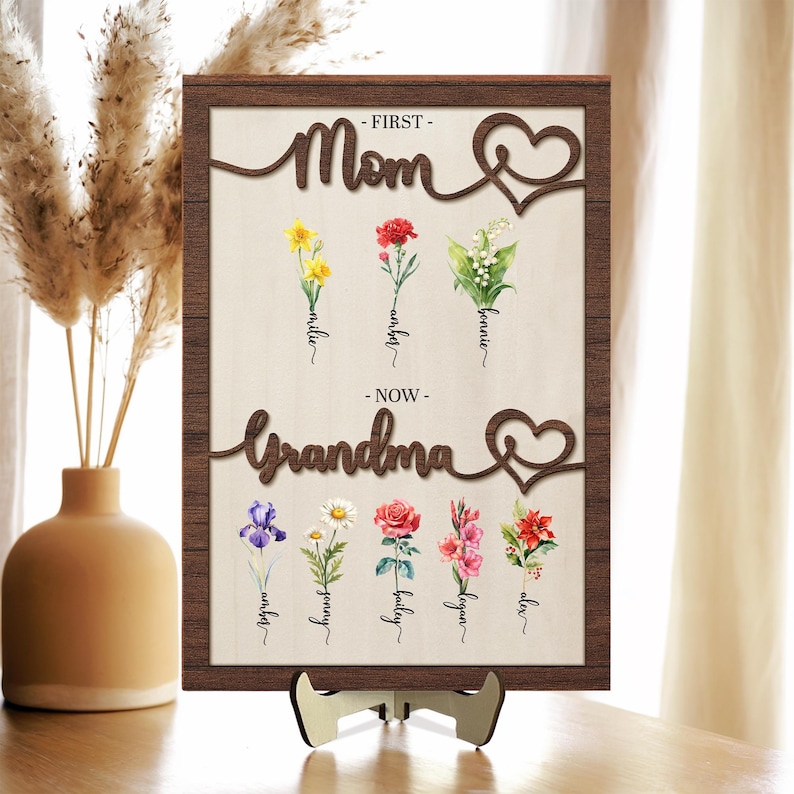 May include: A wooden sign with the words "First Mom" and "Now Grandma" in a cursive font. The sign is decorated with flowers and hearts. The flowers are labeled with names, such as "Millie", "Wyndel", "Bonnie", "Audrey", "Jenny", "Bailey", "Lequan", and "Alex".