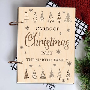 May include: A wooden scrapbook with a light brown finish. The cover features the text "Cards of Christmas Past" and "The Martha Family" along with a decorative design of Christmas trees and snowflakes.