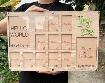 May include: A wooden photo frame with 12 spaces for pictures, labeled with months of the year. The frame has a light wood finish and is decorated with the name "Ivy Ellis" and the text "Happy first birthday!" The frame is designed to hold photos of a baby's first year.