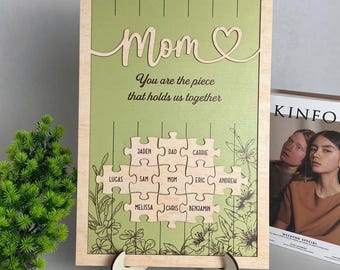 Puede incluir: Un letrero de madera con fondo verde y un corazón recortado. El letrero dice "Mamá" y "Eres la pieza que nos mantiene unidos." Debajo del texto hay un diseño de rompecabezas con nombres como "Jaden", "Papá", "Carrie", "Lucas", "Sam", "Mamá", "Eric", "Andrew", "Melissa", "Chris" y "Benjamin".