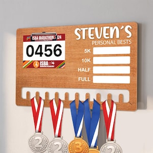 May include: Wooden medal display board with the text "STEVEN'S PERSONAL BESTS". Includes a race bib with the number 0456 and a rack for hanging medals. Several medals with red and blue ribbons are displayed.