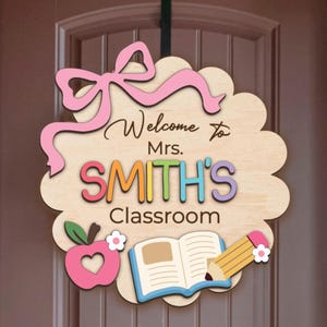 K&ouml;nnte beinhalten: Ein h&ouml;lzernes Willkommensschild f&uuml;r ein Klassenzimmer mit einer rosa Schleife, dem Text "Willkommen in der Klasse von Frau SMITH", einem Apfel, einem Buch und einem Bleistift.