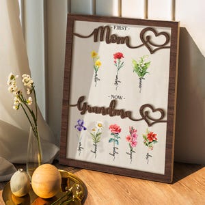 May include: A wooden framed print with the words "First - Mom" and "Now - Grandma" in a decorative font. The print features a heart design and illustrations of flowers with their names written below.