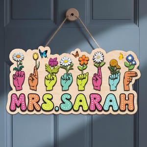 Puede incluir: Cartel de madera con el nombre "MRS. SARAH" en letras coloridas. El cartel presenta manos de varios colores sosteniendo flores, con mariposas. El cartel está colgado de una puerta de madera.