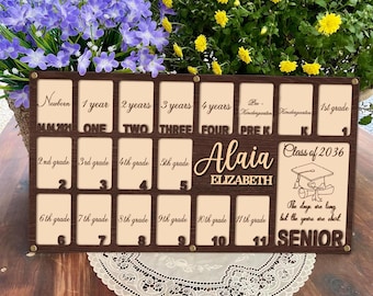 May include: A brown wooden photo frame with 12 slots for pictures. The frame has the words "Newborn" to "11th grade" printed on it. The frame also has the name "Alaia Elizabeth" and the text "Class of 2036" printed on it. The frame is decorated with a graduation cap and the text "The days are long but the years are short" and "Senior".