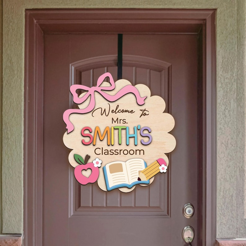 K&ouml;nnte beinhalten: Ein h&ouml;lzernes Willkommensschild mit einer rosa Schleife, dem Text "Welcome to Mrs. SMITH'S Classroom", einem Apfel, einem Buch und einem Bleistift.