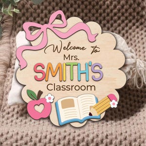 K&ouml;nnte beinhalten: Willkommensschild in Form einer Wolke aus Holz mit einer rosa Schleife, einem Apfel, einem Buch und einem Bleistift. Auf dem Schild steht "Willkommen in der Klasse von Frau SMITH".