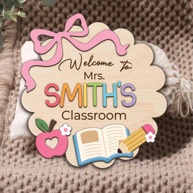 K&ouml;nnte beinhalten: Willkommensschild in Form einer Wolke aus Holz mit einer rosa Schleife, einem Apfel, einem Buch und einem Bleistift. Auf dem Schild steht "Willkommen in der Klasse von Frau SMITH".