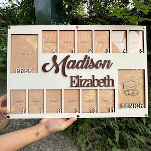 Op de afbeelding: Wit houten fotolijst met 12 fotovakken voor elk schooljaar van de kleuterklas tot en met het laatste jaar. De lijst heeft de naam "Madison Elizabeth" in bruine cursieve letters.