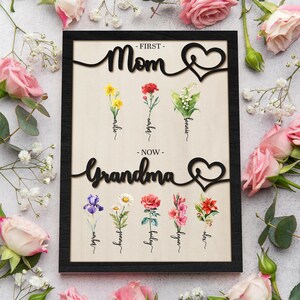 May include: A black framed wood sign with the words "First - Mom - Now - Grandma" in a cursive font. The sign features a heart design and floral illustrations with names written below each flower.