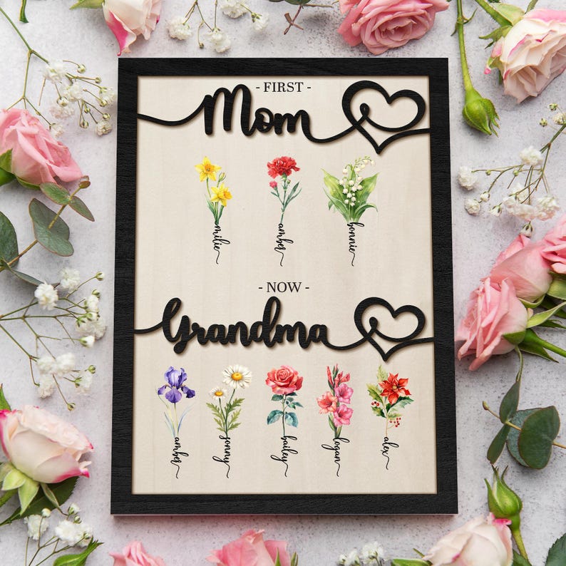 May include: A black framed wood sign with the words "First - Mom - Now - Grandma" in a cursive font. The sign features a heart design and floral illustrations with names written below each flower.