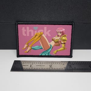 May include: A rectangular patch with a black border featuring an anime-style illustration of a woman in gold armour and a teal skirt, with the word "thick" above. The patch is approximately 7.6 cm wide.