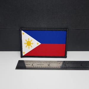 May include: Rectangular patch featuring the flag of the Philippines. The flag design includes a white triangle with a yellow sun and three stars, a blue upper section, and a red lower section. The patch is approximately 3 inches wide.