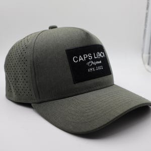 May include: A gray baseball cap with a curved brim. The cap features a perforated side panel and a black square patch with the text "CAPS LOCK Original EST. 2022" in white. The cap is a solid color and has a structured design.