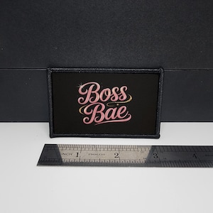 Puede incluir: Parche rectangular negro con las palabras "Boss Bae" en letras rosas con un acento dorado. El parche mide aproximadamente 7,6 cm de ancho y 3,8 cm de alto.