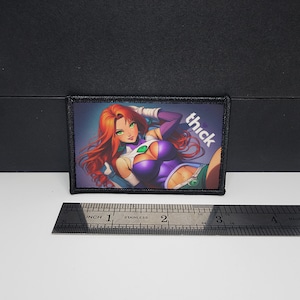 May include: A rectangular patch featuring a comic-style illustration of a woman with red hair, green eyes, and a purple and green outfit. The word "thick" is printed in white. The patch is bordered in black.