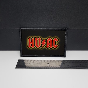 May include: A black rectangular patch featuring the text "HV/AC" in red with a yellow outline. The patch is about 7.6 cm wide. A stainless steel ruler is placed below the patch for scale.