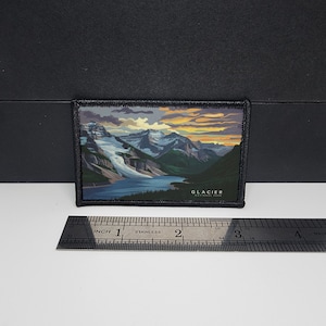 Puede incluir: Parche rectangular con un paisaje del Parque Nacional Glacier. La imagen muestra un lago, montañas y una puesta de sol. El parche tiene un borde negro y el texto "GLACIER NATIONAL PARK". El parche mide aproximadamente 7.6 cm de ancho.