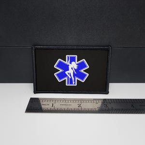 Peut inclure: Écusson rectangulaire noir avec un symbole des services médicaux d'urgence bleu et blanc et une silhouette de femme. L'écusson mesure environ 7,6 cm de large. L'écusson est sur un fond noir.