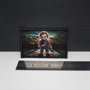 May include: A rectangular patch featuring a menacing cartoon character with red hair and blue eyes, holding a knife. The character is standing on a road with a sunset background. The patch is approximately 3 inches wide.