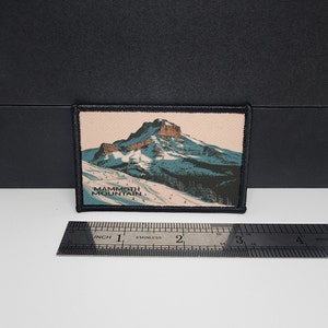 May include: Rectangular patch featuring a mountain landscape with the text "Mammoth Mountain." The patch has a black border and a detailed illustration of a snow-covered mountain with skiers and trees. The patch measures approximately 3 inches in length.