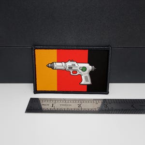 Könnte beinhalten: Rechteckiger Aufnäher mit einem weißen Laserpistolen-Design auf einem Hintergrund aus gelben, roten und schwarzen vertikalen Streifen. Der Aufnäher hat einen schwarz genähten Rand. Ein Edelstahl-Lineal ist darunter platziert.
