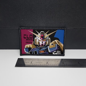 May include: A rectangular patch featuring a detailed illustration of a robot's head and upper body, with gold, white, blue, and red accents. The patch includes the text "CLO" and "0026". A stainless steel ruler is placed below the patch.