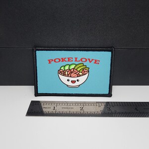 May include: A rectangular patch with a light blue background and black border. The patch features the words "POKE LOVE" in red above a cartoon bowl of poke. A stainless steel ruler is placed below the patch, measuring approximately 3 inches.