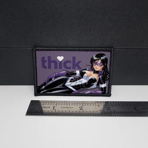 Peut inclure: Un écusson rectangulaire avec le mot "thick" en lettres blanches et un symbole de cœur. L'écusson présente une illustration de style bande dessinée d'une femme en costume de super-héros noir et blanc, avec des accents violets et un masque. L'écusson mesure environ 7.6 cm de large.