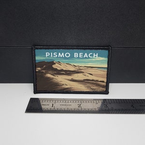 Op de afbeelding: Rechthoekige patch met een landschap van Pismo Beach met zandduinen onder een blauwe lucht. De tekst "PISMO BEACH" staat bovenaan. De patch is omrand met zwarte stiksels. De lengte is ongeveer 7.6 cm.