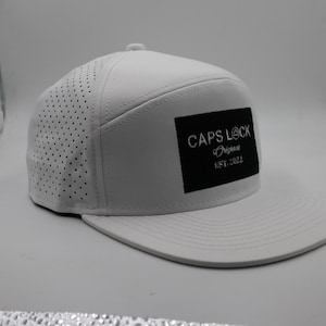 Peut inclure: Casquette de baseball blanche avec une visière plate. Le chapeau présente un design perforé sur les panneaux latéraux et un patch rectangulaire noir avec le texte "CAPS LOCK Original EST. 2022".