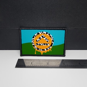Puede incluir: Parche rectangular con borde negro. El parche presenta un fondo de cielo azul y hierba verde. Un círculo con un borde de estampado de vaca rodea un triángulo naranja. Goteos naranjas caen de la parte inferior del círculo.