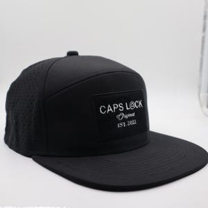May include: Black flat-brimmed baseball cap with a perforated side panel. The cap features a square patch with the text "CAPS LOCK Original EST. 2022" in white. The hat has a structured crown and a flat brim.