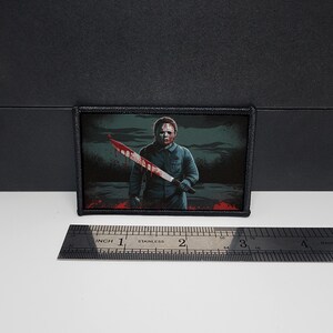 May include: Rectangular patch featuring a horror movie character holding a bloody machete. The patch has a black border and a dark background with red splatters. The image measures approximately 3 inches in length.