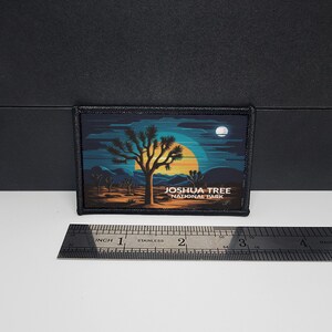 Op de afbeelding: Een rechthoekige patch met een woestijnlandschap met een Joshua-boom tegen een zonsondergang. De patch bevat de tekst "Joshua Tree National Park". De patch is omrand met zwarte stiksels.